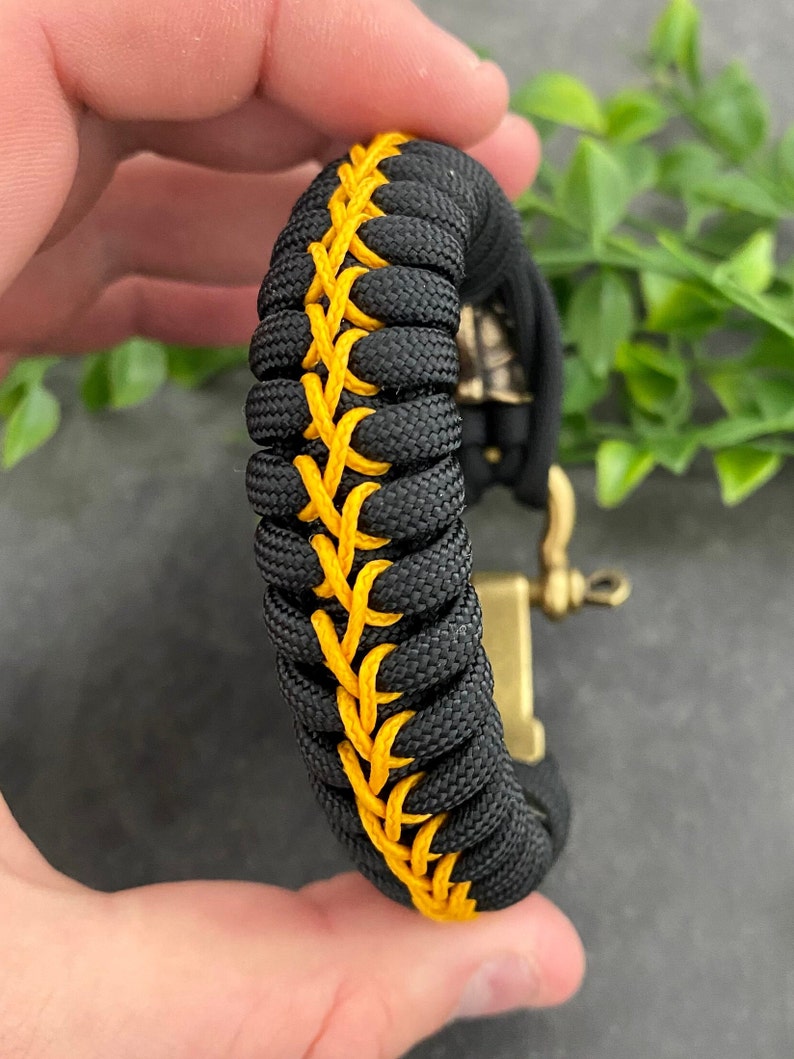 May include: Black paracord bracelet with a yellow stitching detail. The bracelet has a metal clasp.