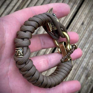 May include: A brown paracord bracelet with a brass clasp and a decorative brass skull charm. The bracelet has a small brass band with a geometric design.