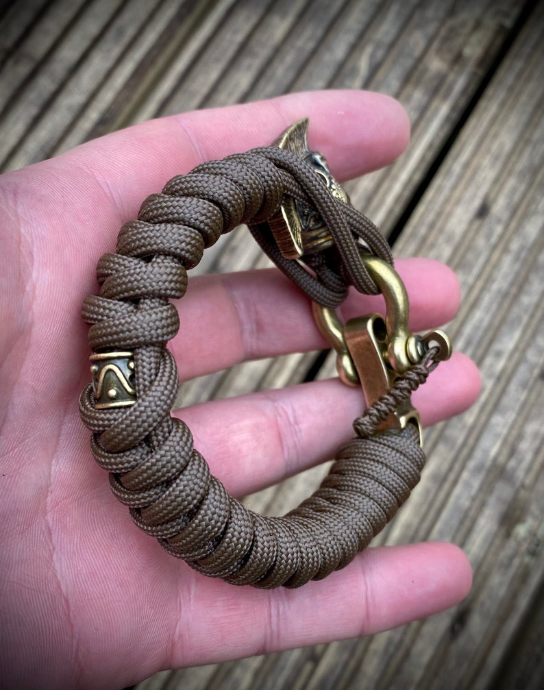 May include: A brown paracord bracelet with a brass clasp and a decorative brass skull charm. The bracelet has a small brass band with a geometric design.