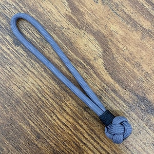 Op de afbeelding: Een grijze paracord sleutelhanger met een zwarte knoop aan het einde.