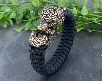 Puede incluir: Un brazalete de paracord negro con un cierre de cabeza de lobo dorado. La cabeza de lobo está detallada con intrincados tallados y tiene una superficie texturizada.
