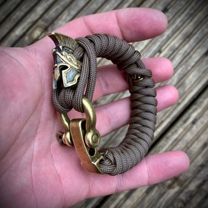 May include: A brown paracord bracelet with a brass clasp and a brass Roman helmet charm.