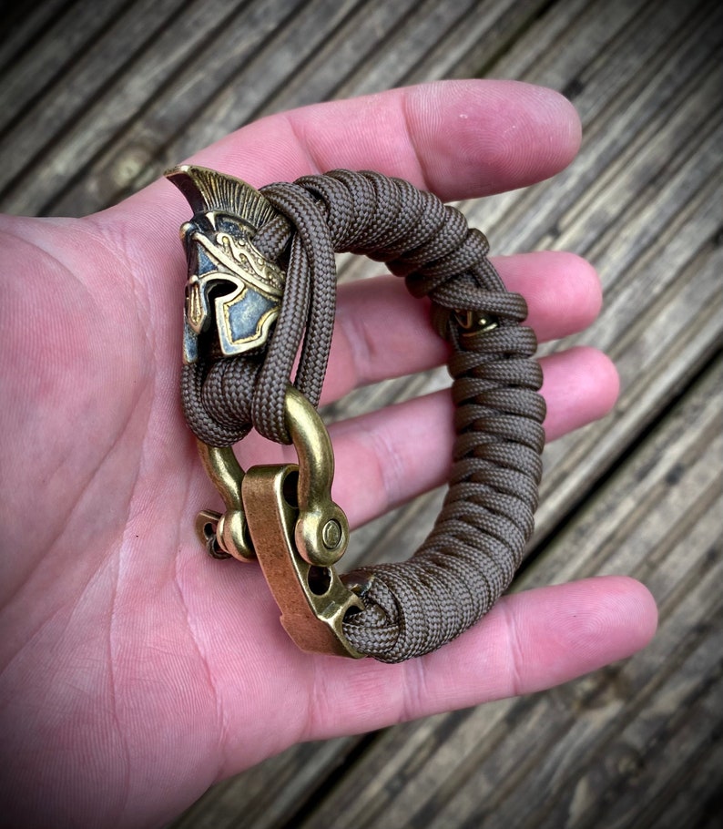 May include: A brown paracord bracelet with a brass clasp and a brass Roman helmet charm.
