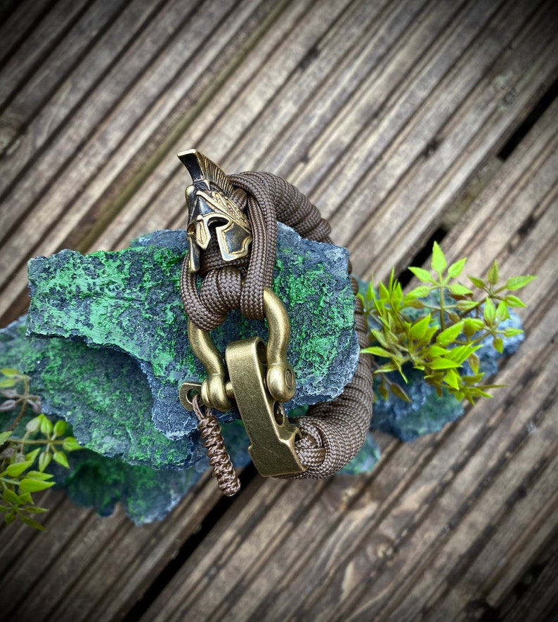 May include: A brown paracord bracelet with a brass clasp and a gold-colored helmet charm. The bracelet is on a green and gray rock.