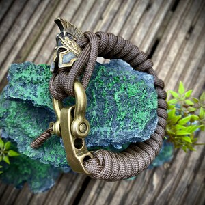 May include: A brown paracord bracelet with a brass clasp and a decorative brass Roman helmet charm.