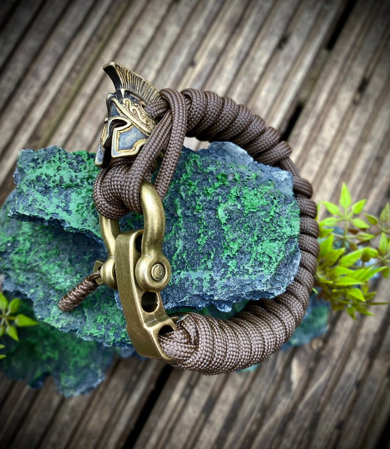 May include: A brown paracord bracelet with a brass clasp and a decorative brass Roman helmet charm.