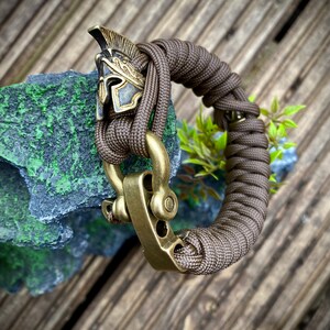 May include: A brown paracord bracelet with a brass clasp and a gold-colored Roman helmet charm.