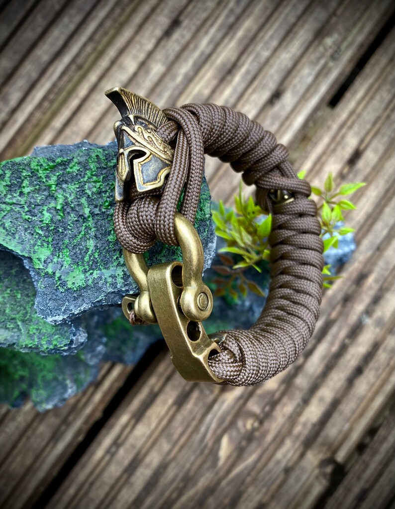 May include: A brown paracord bracelet with a brass clasp and a gold-colored Roman helmet charm.