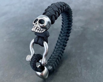 May include: Black paracord bracelet with a silver skull charm and a silver clasp. The bracelet is made of a braided paracord and has a skull charm on one end and a clasp on the other.