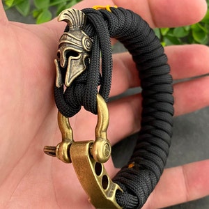 May include: Black paracord bracelet with a gold-toned metal clasp and a small, detailed, gold-toned metal helmet charm.