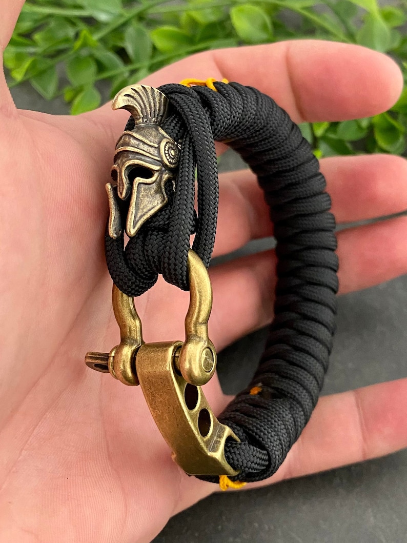 May include: Black paracord bracelet with a gold-toned metal clasp and a small, detailed, gold-toned metal helmet charm.