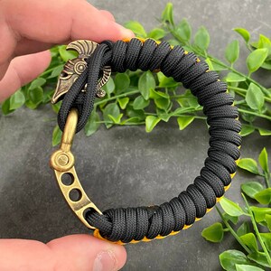 May include: Black paracord bracelet with a yellow accent and a brass clasp featuring a helmet design.