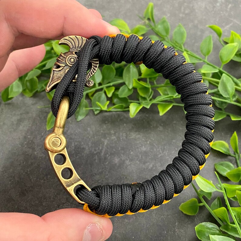 May include: Black paracord bracelet with a yellow accent and a brass clasp featuring a helmet design.