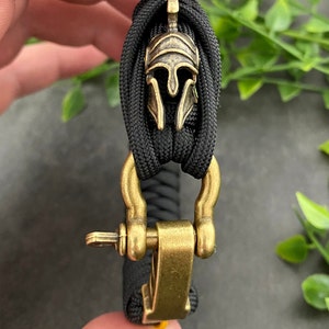 May include: A black paracord bracelet with a gold metal clasp and a gold metal Spartan helmet charm.
