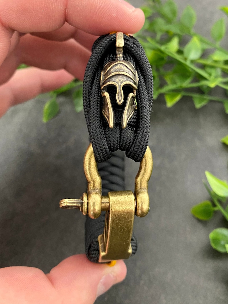 May include: A black paracord bracelet with a gold metal clasp and a gold metal Spartan helmet charm.