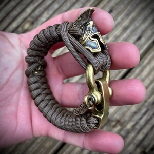 May include: A brown paracord bracelet with a brass clasp and a gold-colored helmet charm.
