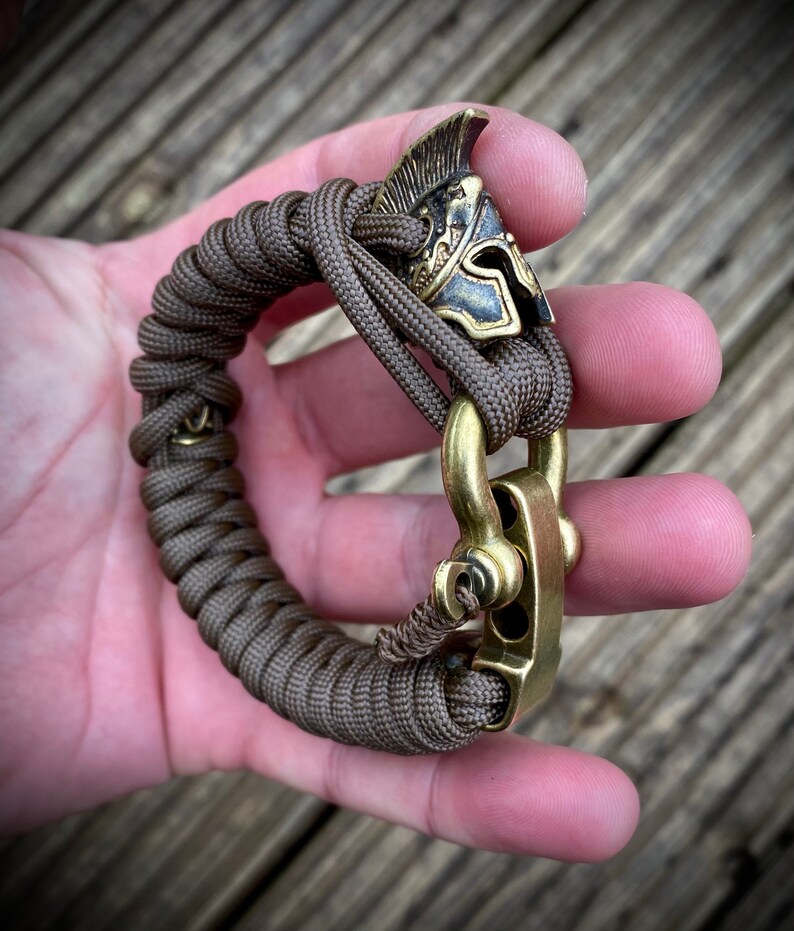 May include: A brown paracord bracelet with a brass clasp and a gold-colored helmet charm.