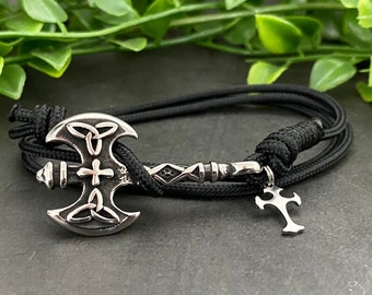 May include: A black paracord bracelet with a silver axe charm and a silver cross charm. The axe charm features a Celtic knot design.