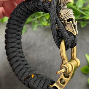 May include: Black paracord bracelet with a gold-toned metal clasp and a small, detailed, gold-toned helmet charm.