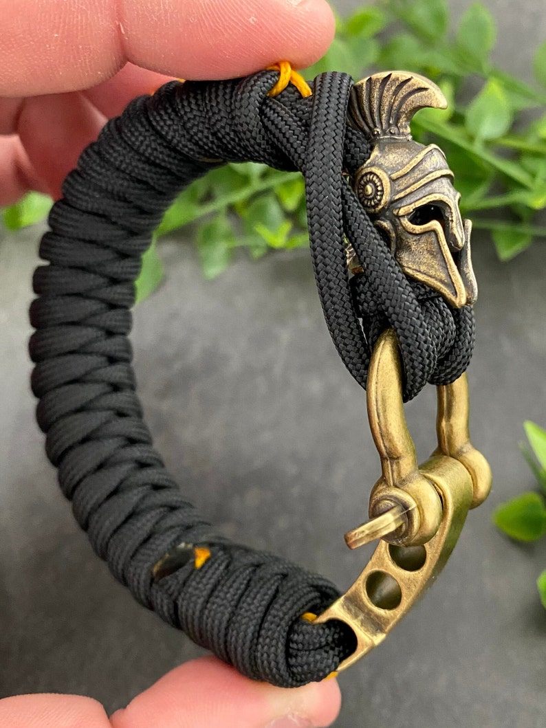 May include: Black paracord bracelet with a gold-toned metal clasp and a small, detailed, gold-toned helmet charm.