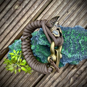 May include: A brown paracord bracelet with a brass clasp and a brass Roman helmet charm.