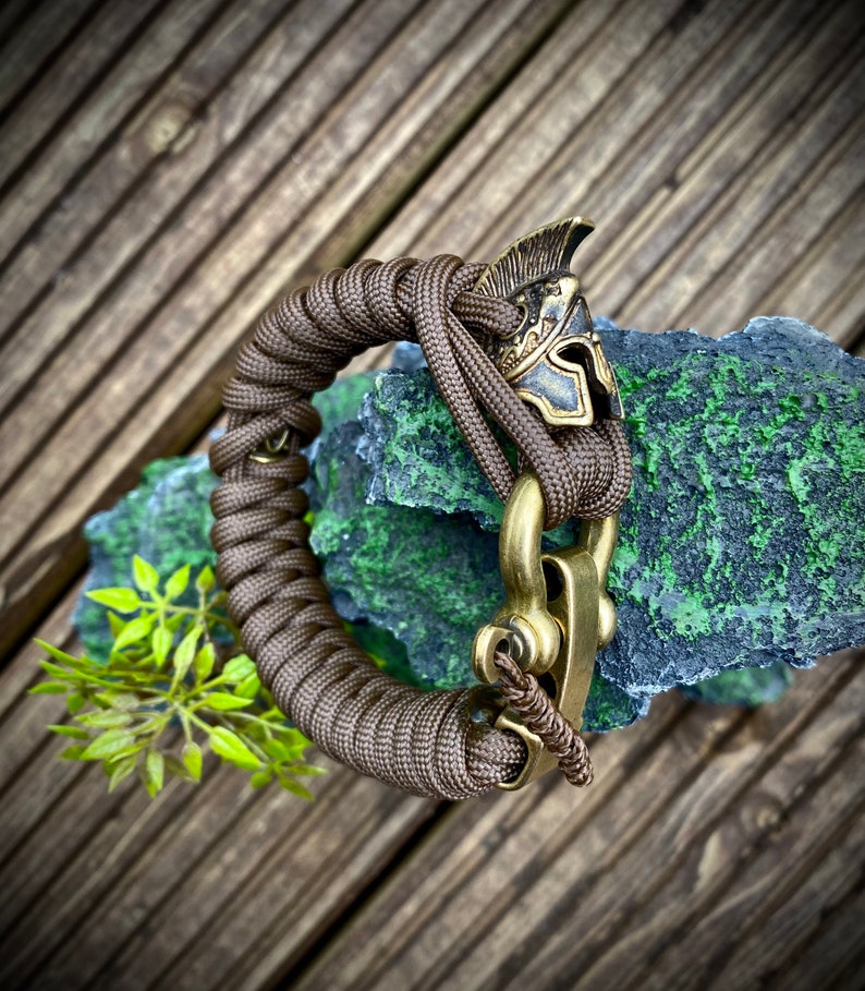 May include: A brown paracord bracelet with a brass clasp and a brass Roman helmet charm.