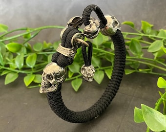 May include: A black paracord bracelet with silver skull charms. The bracelet is made of a thick, durable cord and features several skull-shaped beads.