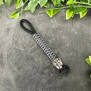 Puede incluir: Un llavero de paracord negro y gris con una cuenta de metal plateado. La cuenta tiene un diseño geométrico.