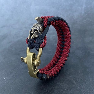 May include: A black and red paracord bracelet with a brass clasp and a small, detailed, silver-coloured helmet charm.