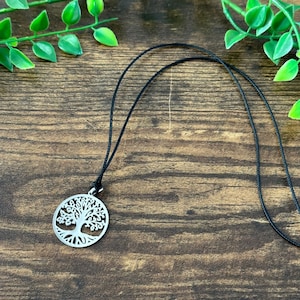 Puede incluir: Un collar con un colgante de plata en forma de árbol de la vida en un cordón negro. El colgante es redondo y presenta un diseño de árbol detallado con ramas y hojas.