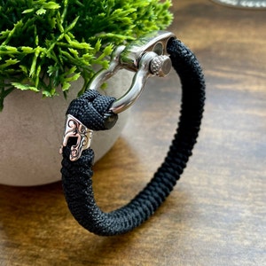 May include: A black paracord bracelet with a silver clasp and a silver decorative charm. The charm features a stylized design.