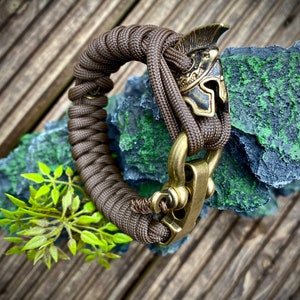 May include: A brown paracord bracelet with a brass clasp and a brass helmet charm.