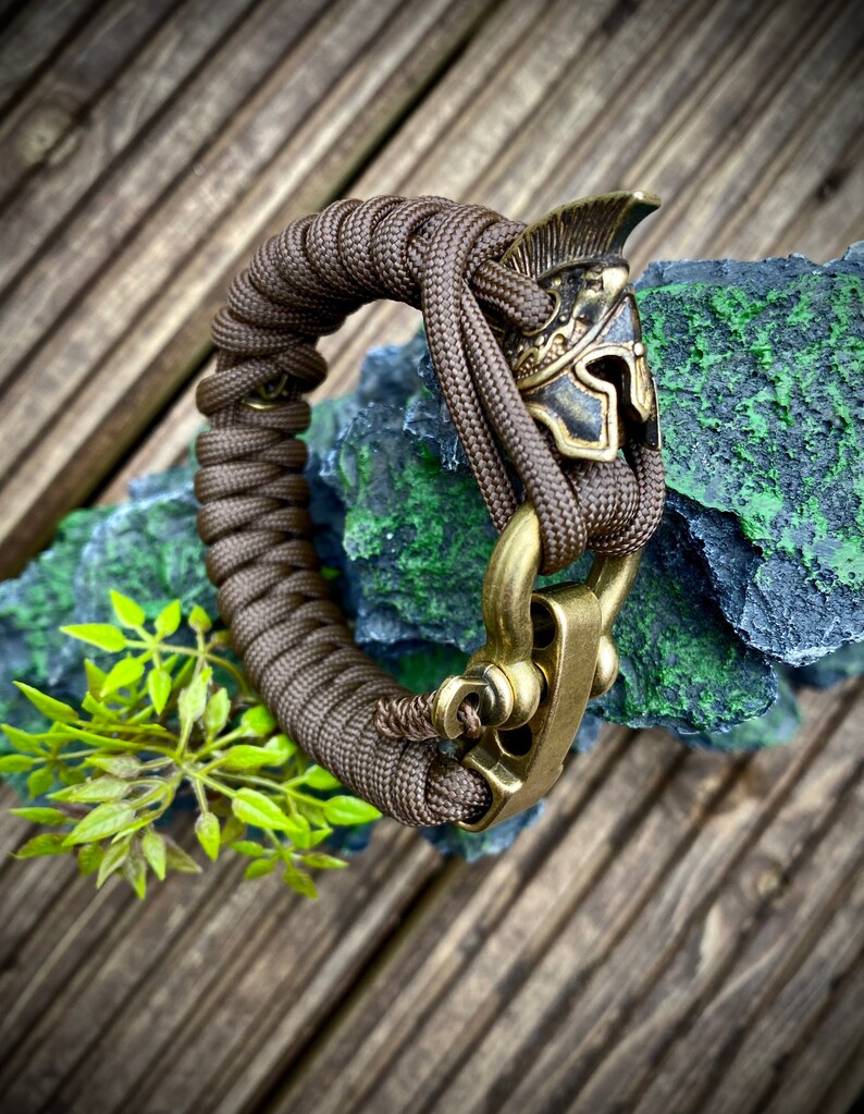 May include: A brown paracord bracelet with a brass clasp and a brass helmet charm.