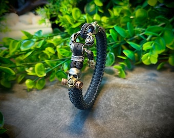 May include: Black braided bracelet featuring skull and gas mask accents. The bracelet has silver and gold-toned metal details. The clasp has a skull design. The bracelet is set against a backdrop of green foliage.