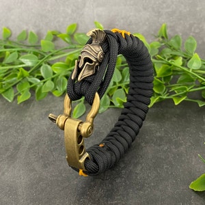 May include: Black paracord bracelet with a gold-toned metal clasp and a small, detailed, gold-toned metal helmet charm.