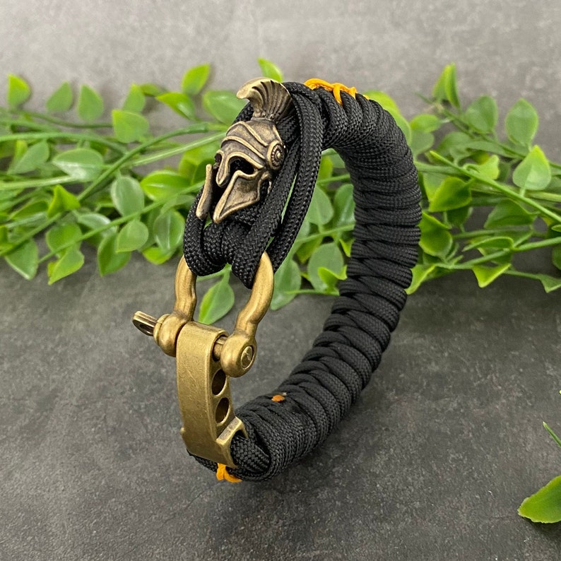 May include: Black paracord bracelet with a gold-toned metal clasp and a small, detailed, gold-toned metal helmet charm.