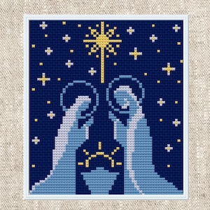 May include: A cross-stitch pattern featuring a nativity scene against a dark blue background. Two figures in light blue robes with halos stand over a manger, illuminated by a large yellow star above and smaller yellow and white stars.