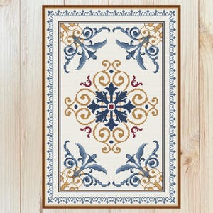 May include: A framed cross-stitch design with a central floral pattern in navy blue, gold, and red on a cream backdrop. The design incorporates elaborate scrollwork and leaf motifs, enclosed by a border of repeating geometric shapes.