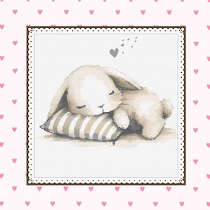 May include: A cross-stitch image of a sleeping bunny on a striped pillow. The bunny is light beige, with closed eyes, and is resting on a gray and white striped pillow. A gray heart and small gray shapes float above the bunny. The background is pink with small pink hearts.
