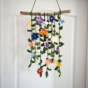 May include: A crocheted wall hanging featuring a variety of colorful flowers and green leaves. The flowers are in shades of purple, blue, orange, yellow, and pink. The hanging is attached to a natural branch.