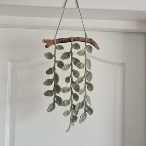 May include: A handmade, crochet wall hanging featuring cascading green leaves. The leaves are suspended from a natural wooden branch, which is attached to a gray cord for hanging. The piece is designed to add a touch of nature-inspired decor to any space.