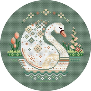 May include: A round, cross-stitched design featuring a white swan with a peach beak and accents, floating on water. The swan is surrounded by peach and green floral elements, set against a muted green background.