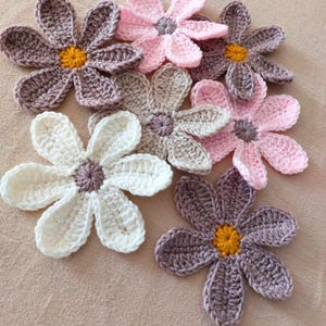 May include: A collection of handmade crochet flowers in various colors, including white, pink, and shades of gray. Each flower features a central yellow or gray accent, with petals arranged around the center. The flowers are displayed on a neutral-toned surface.