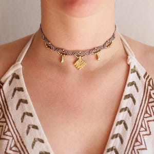 Può includere: Un girocollo in macramé grigio con perline dorate e un ciondolo dorato. Il ciondolo è un quadrato con un design geometrico.