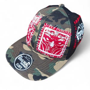 Könnte beinhalten: Snapback-Cap mit Camouflage-Print und rot-weißen Patches mit einem grafischen Gesichtsdesign. Die Kappe hat eine schwarze Mesh-Rückseite und einen gebogenen Schirm. Das Cap enthält ein rundes Etikett mit der Aufschrift "THE ORIGINAL SNAPBACK ATLANTIS."