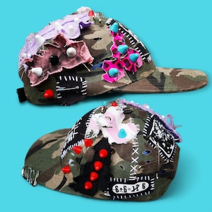 Könnte beinhalten: Zwei Camouflage-Baseballkappen, verziert mit Stoffflicken, Nieten und Sicherheitsnadeln. Eine Kappe hat einen pinkfarbenen Leopardenmuster-Akzent, die andere zeigt den Text "6+6=12". Beide Kappen haben einen schwarzen Schirm, vor einem hellblauen Hintergrund.