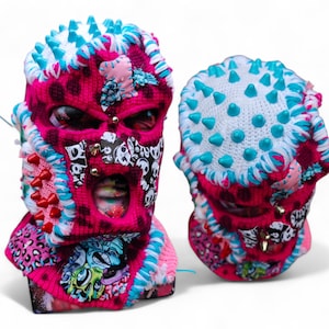 Pode incluir: Duas balaclavas feitas à mão. A balaclava principal é rosa choque com bolinhas pretas, adornada com detalhes brancos, azuis e rosa. Uma balaclava tem um design de caveira e ossos cruzados. Ambas têm pontas e tachas.
