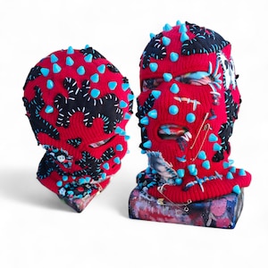 Könnte beinhalten: Zwei rote Sturmhauben mit schwarz-weißen Akzenten und hellblauen Stacheln. Eine Sturmhaube hat Sicherheitsnadeln und Patches. Die Sturmhauben werden auf einer farbenfrohen, strukturierten Oberfläche präsentiert. Einzigartiges, künstlerisches Design.