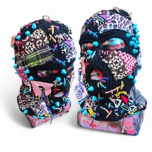 May include: Two black balaclavas with colourful fabric patches, blue spikes, and safety pins. The balaclavas have eye and mouth openings. One has a pink butterfly charm and the other has a purple charm. The balaclavas are displayed on a painted surface.
