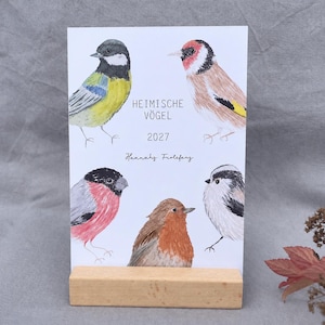 Puede incluir: Una tarjeta de calendario 2027 titulada HEIMISCHE V&Ouml;GEL con ilustraciones en acuarela de cinco aves, incluyendo un carbonero com&uacute;n, un jilguero, un camachuelo, un petirrojo y un mito, exhibida sobre un soporte de bloque de madera clara.